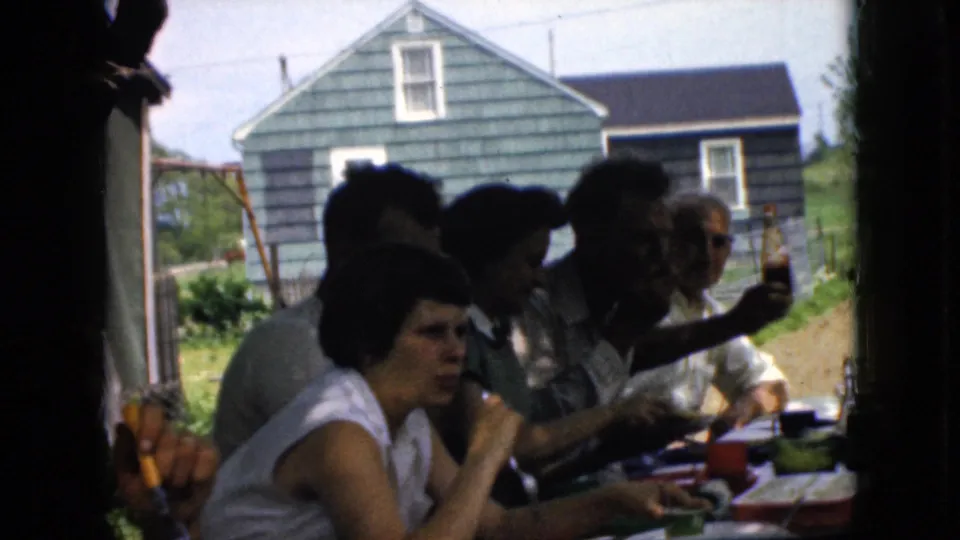 1969 California home-movie frame of an outdoor family meal, with adults partly silhouetted at the table and one man holding up a bottle toward the camera.