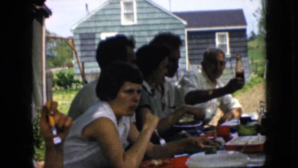 1969 California home-movie frame showing a family group seated outdoors at a meal, with a woman in front, men and women behind her, and a green-sided house across the yard.