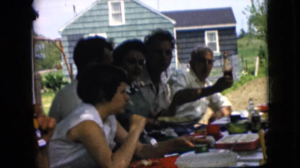 1969 California home-movie frame of adults gathered around a backyard table, with faces in shadow, food and tableware in front, and a man lifting a bottle near the house behind them.