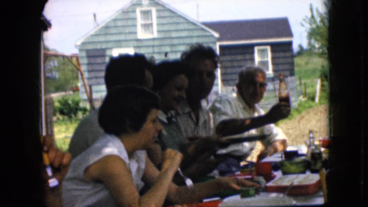 Adults sit around an outdoor table with food containers and bottles, seen through a dark window frame.