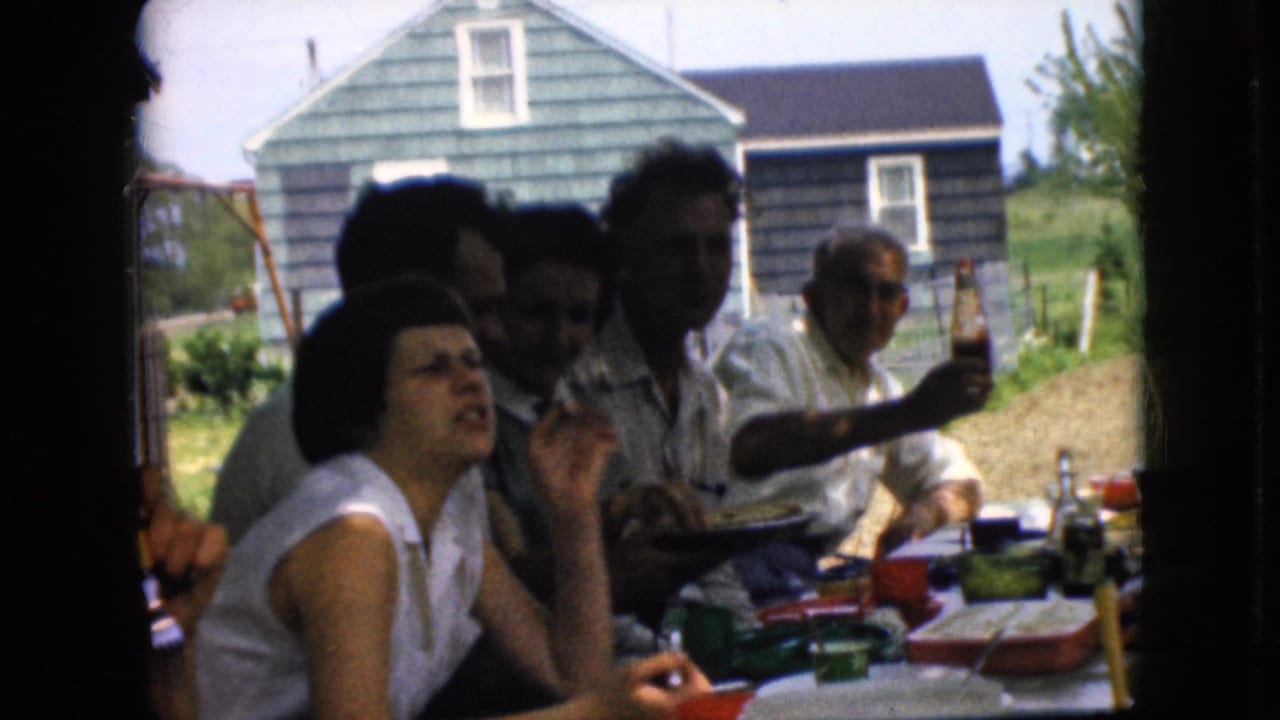 A woman in a white sleeveless top sits at the table while several adults gather behind her outdoors.