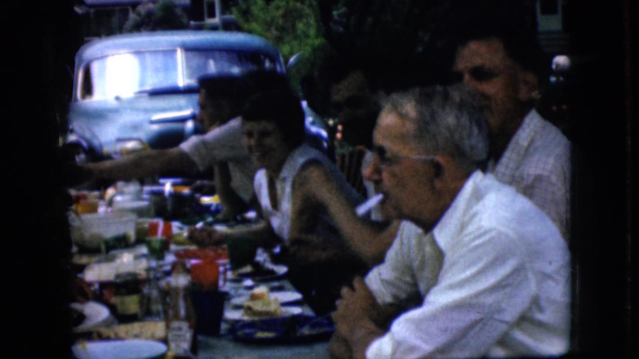 In supported 1969 California home-movie footage, older adults sit closely at a shaded outdoor picnic table while eating and smoking, with a light blue older vehicle visible in the background.