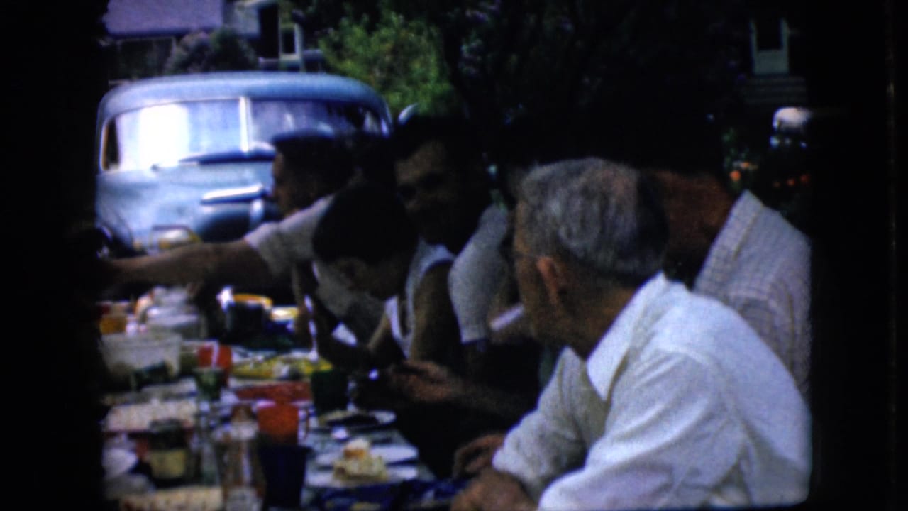 Older adults at a shaded outdoor picnic table in supported 1969 California footage eat and converse as one person reaches across the table, with the light blue vehicle still visible behind them.