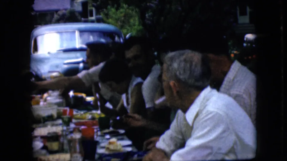 Older adults at a shaded outdoor picnic table in supported 1969 California footage eat and converse as one person reaches across the table, with the light blue vehicle still visible behind them.