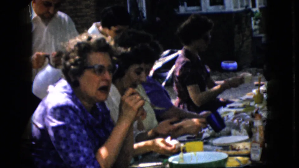 The home-movie frame shows older adults, including women in patterned clothing, seated at an outdoor table with plates, cups, bottles, and food in a residential-looking setting.