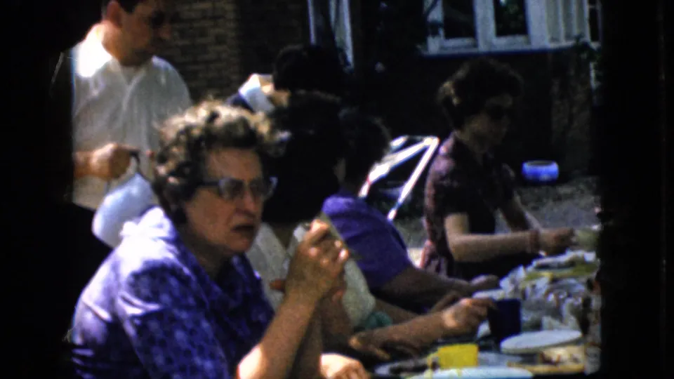 In the supported 1969 California picnic scene, a woman with glasses sits near the camera holding food or a utensil while other adults continue eating at the crowded outdoor table.