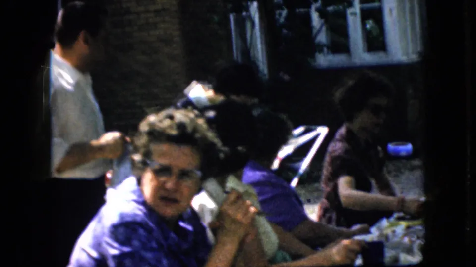 1969 California 8mm frame showing women seated at a long outdoor brunch table with plates and cups while a man in a white shirt stands in the background.