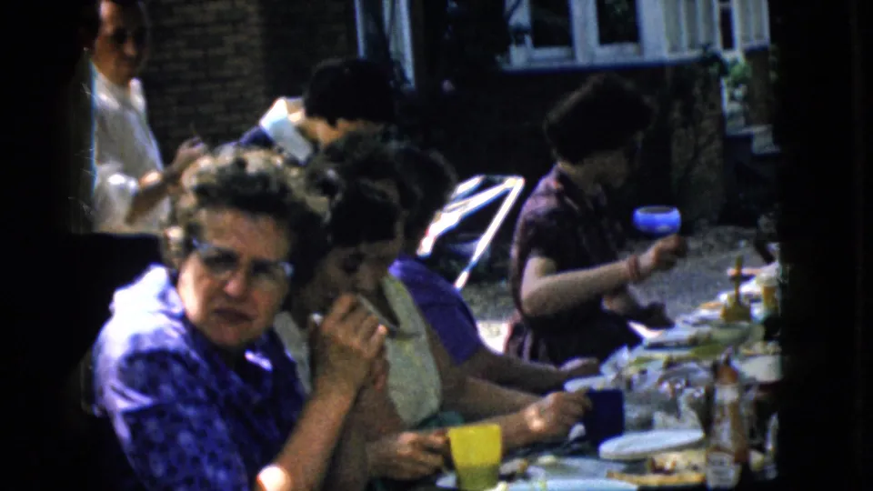 1969 California home-movie frame with a woman in glasses and a blue patterned top looking toward the camera as other women eat at the outdoor table.