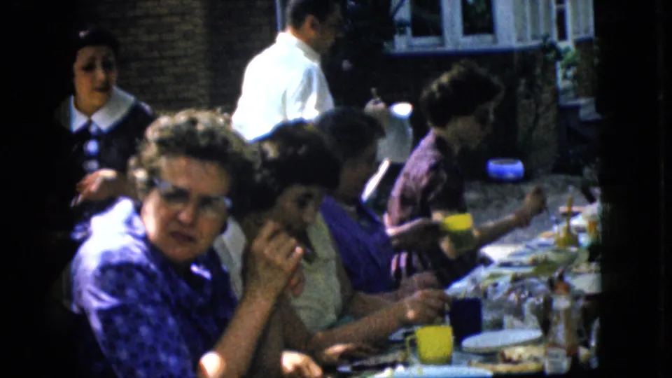 1969 California 8mm frame showing several women at an outdoor brunch table, with a dark-clothed figure at left and a man in white moving behind them.