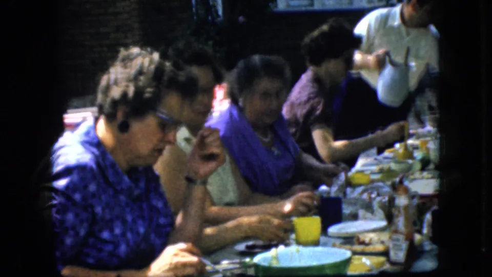 1969 California home-movie frame showing women seated closely at a long outdoor table with plates, cups, and a person serving from behind.