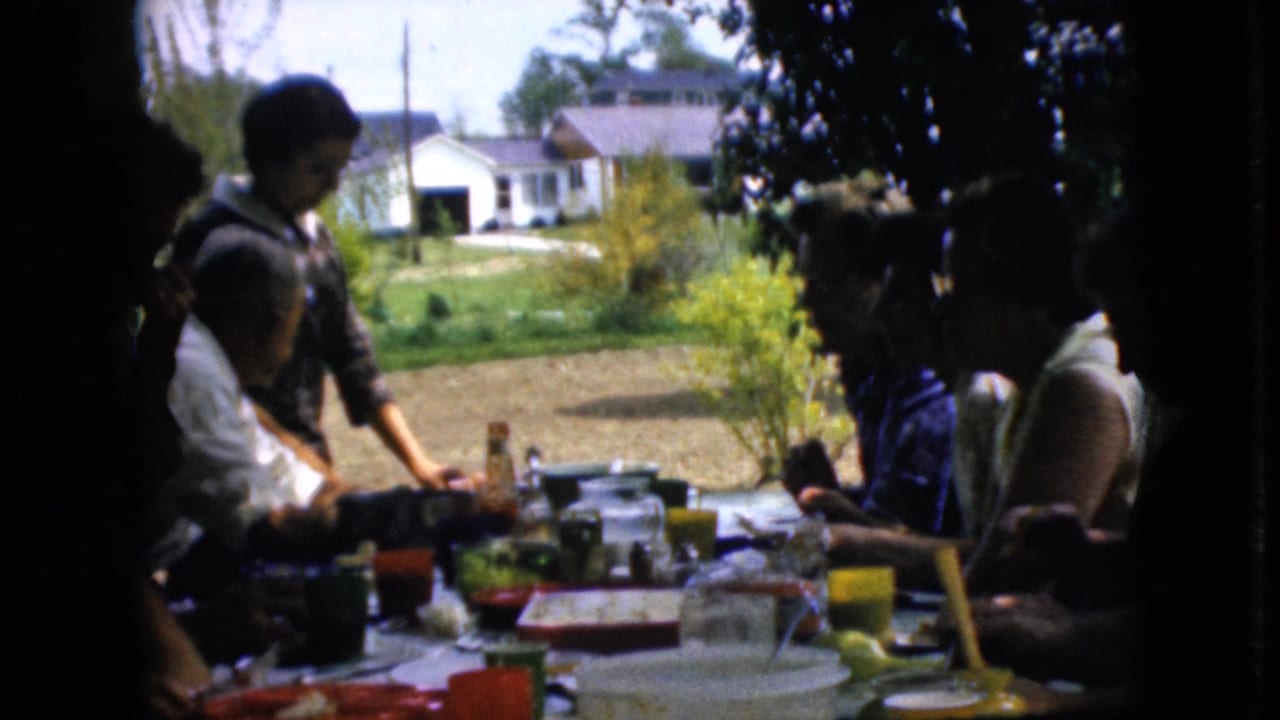 1969 California home-movie frame showing family members around a long outdoor picnic table with food, bright cups, lawn, shrubs, and a white house beyond.