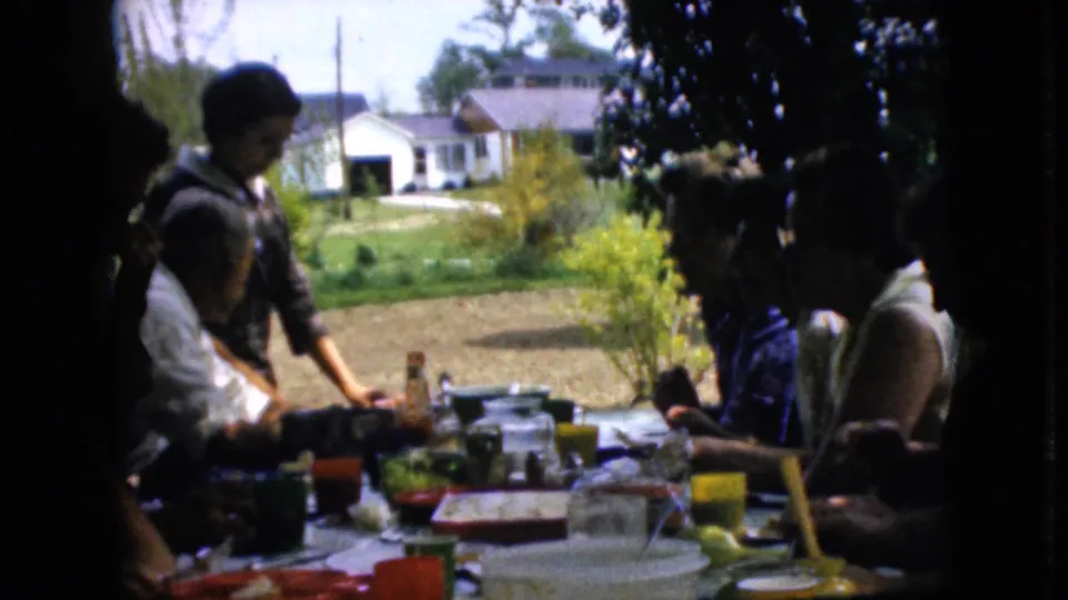 1969 California home-movie frame showing family members around a long outdoor picnic table with food, bright cups, lawn, shrubs, and a white house beyond.