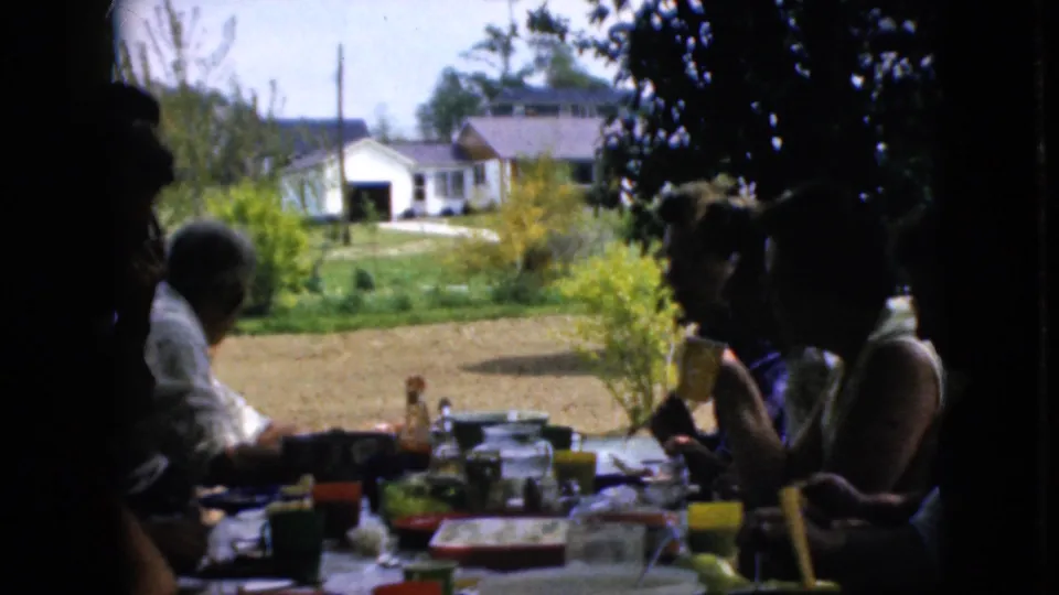 1969 California 8mm frame of adults gathered at an outdoor meal, partly silhouetted by shade, with picnic table items and a residential yard visible behind them.