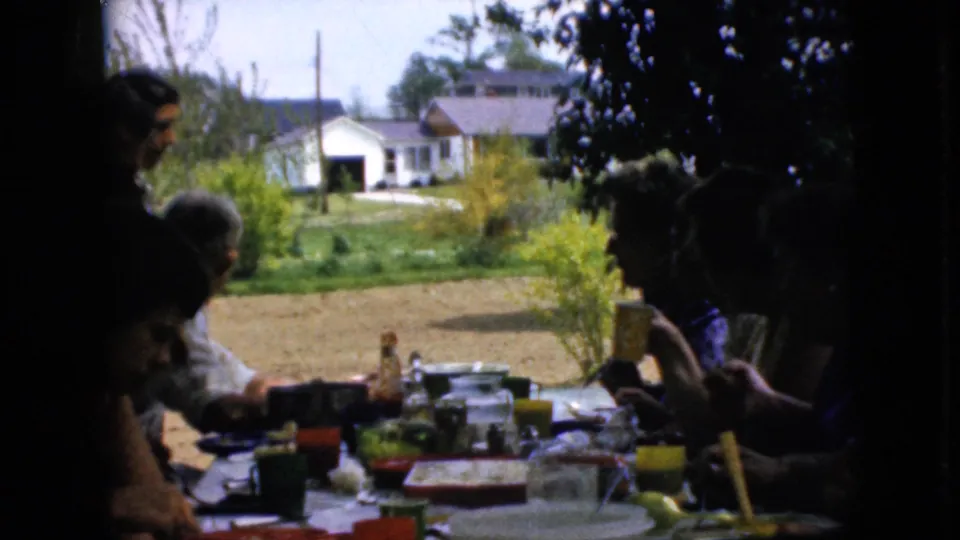1969 California home-movie frame showing a candid family gathering at a picnic table under shade, with cups, dishes, shrubs, lawn, and a white house in the distance.