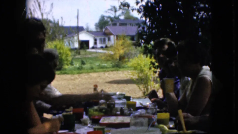 1969 California 8mm frame of people seated at a backyard picnic table, one holding a yellow cup while others reach among dishes and cups, with a lawn and house behind.