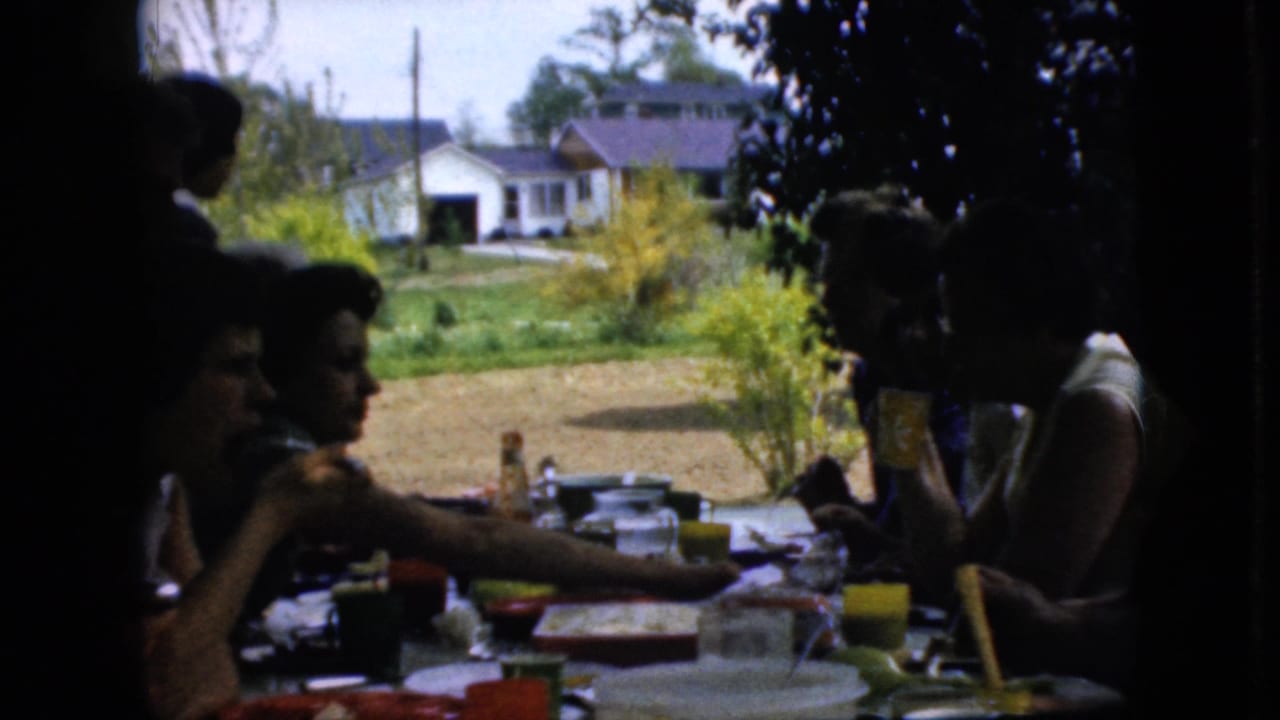 1969 California home-movie frame showing adults at a shaded outdoor family table with dishes, cups, bottles, trees, lawn, and neighboring houses visible in the background.
