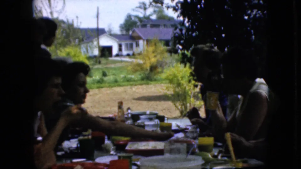 1969 California home-movie frame showing adults at a shaded outdoor family table with dishes, cups, bottles, trees, lawn, and neighboring houses visible in the background.