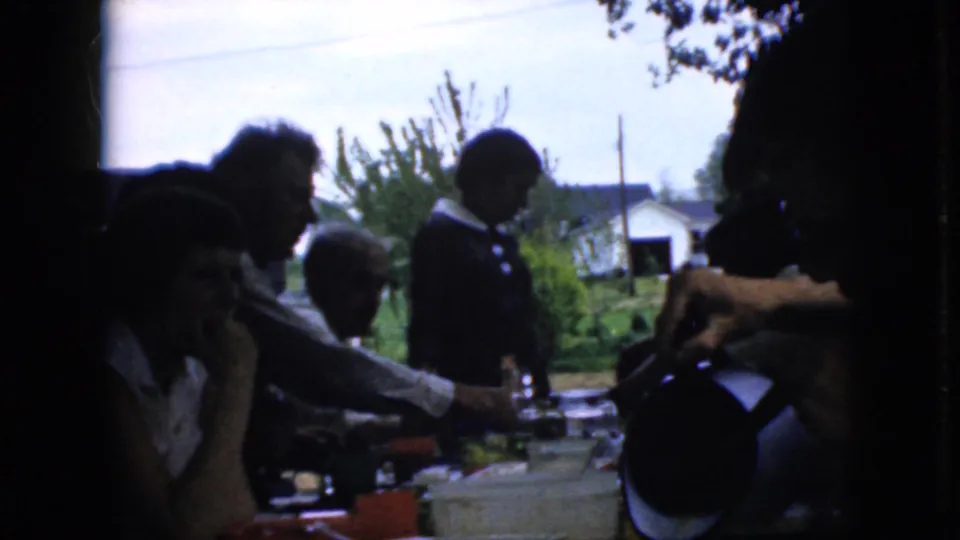 1969 California home-movie frame showing family members seated at a crowded outdoor table as someone stands nearby and another person reaches across the dishes and drinks.
