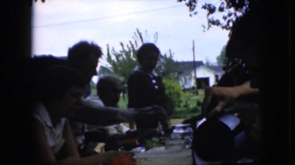 1969 California home-movie frame of an outdoor family meal with adults in shade, a person standing by the table, visible dishes and cups, and suburban greenery behind them.