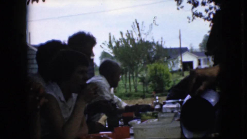 A 1969 California home-movie frame shows family members in silhouette at an outdoor meal, with a serving gesture near a pitcher and suburban houses in the background.