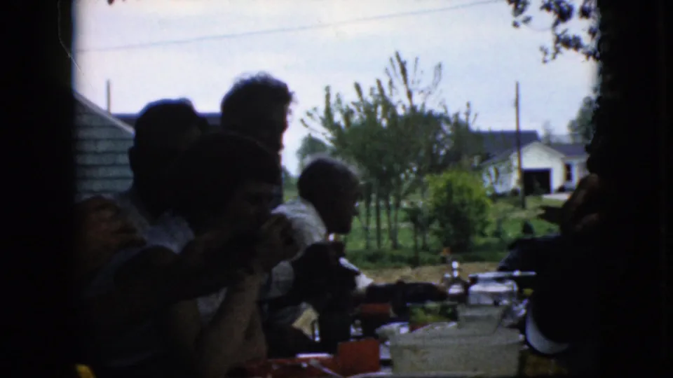 The shaded 8mm frame shows a family meal outdoors in California in 1969, with seated figures, tableware, garden plants, and nearby houses visible beyond the table.