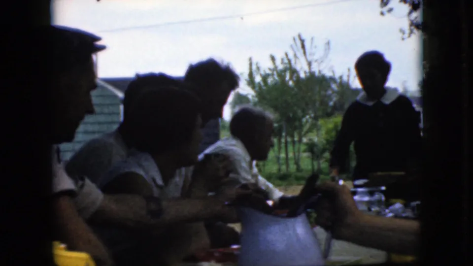 A standing figure appears beside seated relatives at a shaded outdoor table in this 1969 California home-movie view, with a pitcher, tableware, trees, and houses behind them.