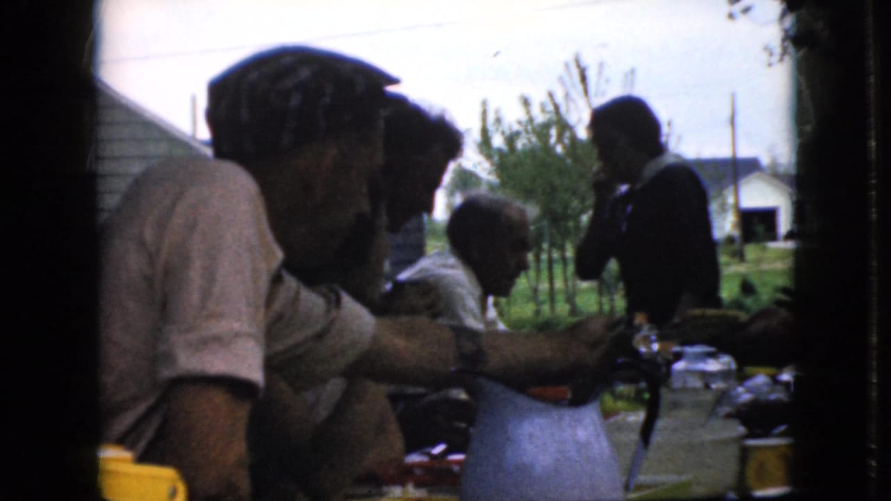 California 1969 home-movie frame showing adults seated around a long outdoor meal table, with food, bottles, and a pitcher visible against a bright suburban yard.