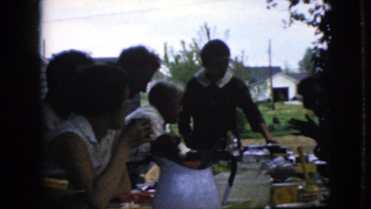 Several people sit and stand around a food-covered outdoor table in a yard.