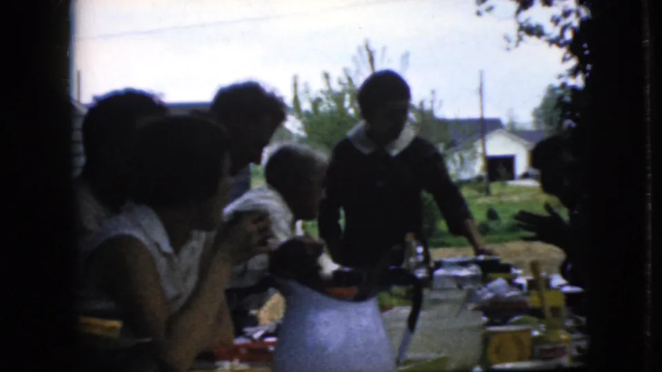 California 1969 8mm frame of people gathered at an outdoor dining table, including a standing young person near the center and seated adults in silhouette.