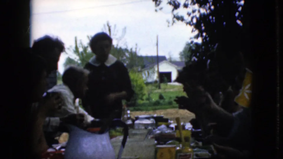 A late-1960s California home-movie view shows seated and standing family members around a crowded outdoor table, with heavy shadow in the foreground and a bright yard beyond.