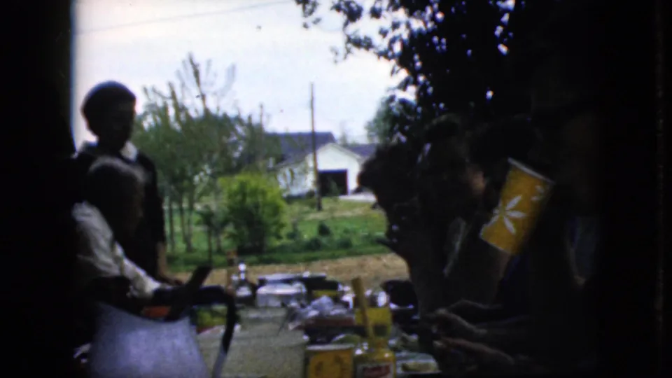 California 1969 home-movie frame with silhouetted diners at an outdoor table, a visible orange or yellow cup near the right edge, and a suburban yard in the background.