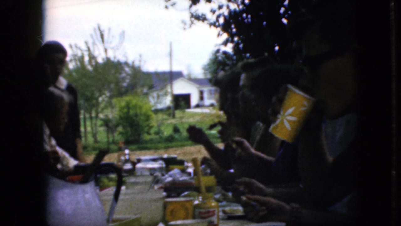 Family members sit and stand around an outdoor table set with food, cups, and a blue pitcher.