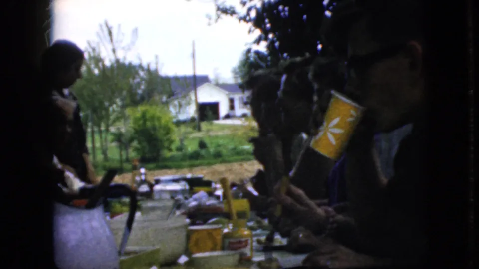A close porch-table view shows a person holding a yellow patterned paper cup while family members sit nearby, with jars, dishes, trees, and a light-colored garage or house in the background.