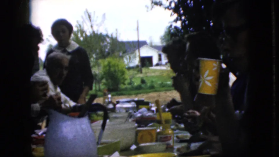 Several family members are visible around the shaded outdoor table, including an older seated man and a standing woman, with a blue pitcher, cups, jars, and a suburban yard behind them.