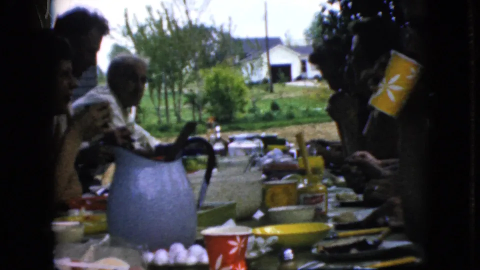 The 1969 California home-movie frame shows relatives seated and standing at a porch lunch table with a blue pitcher, eggs or small round foods, patterned cups, and greenery in the background.