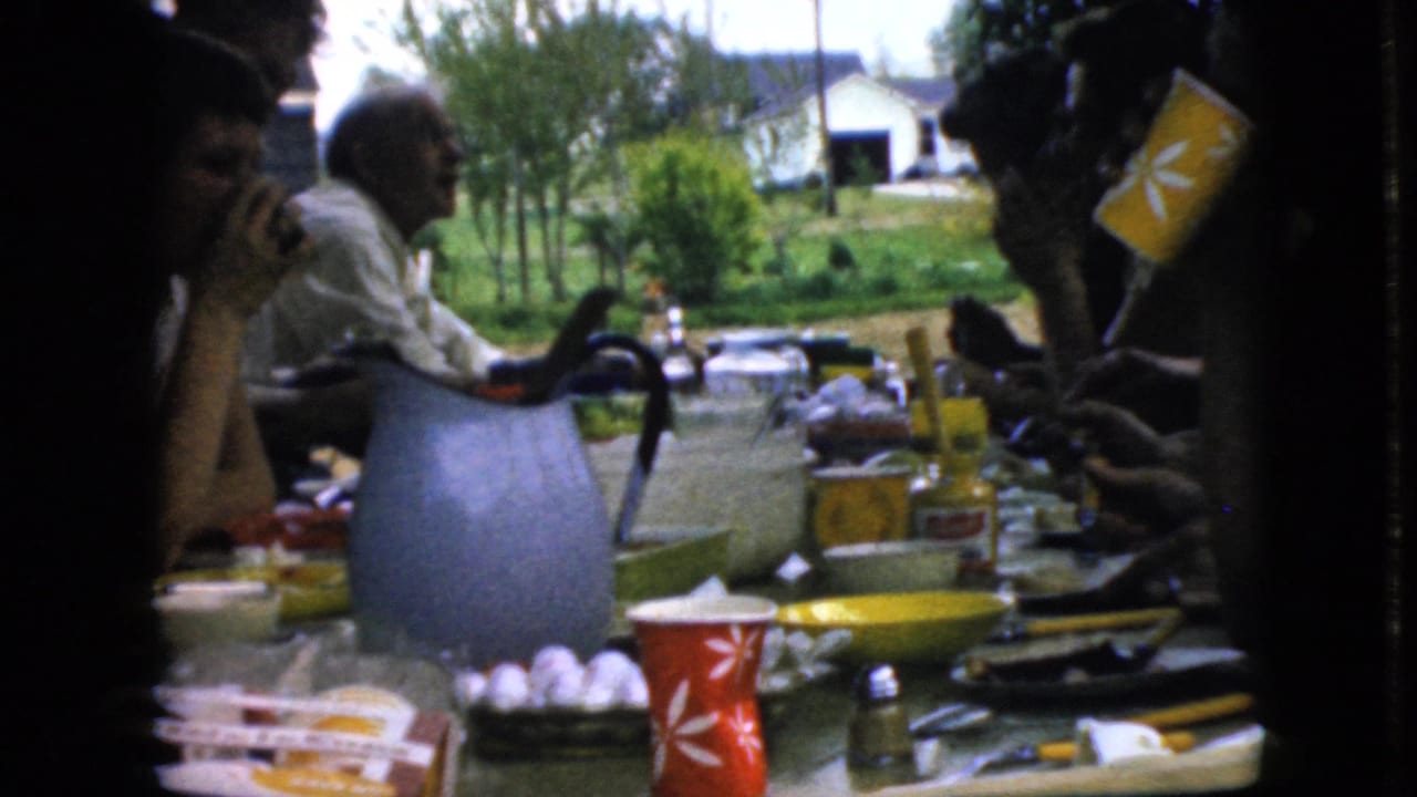 People seated at an outdoor table with a blue pitcher, cups, and food in front of a grassy yard.