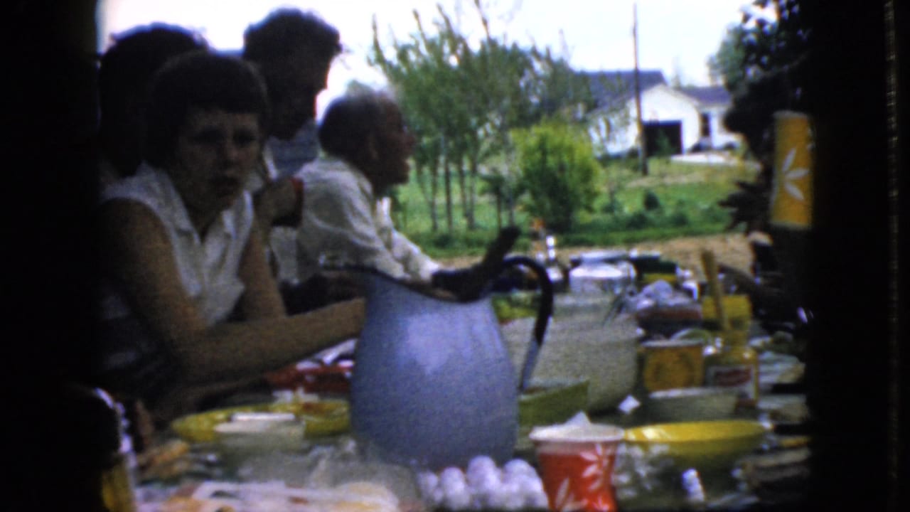 A young person and adults sit at an outdoor table with a blue pitcher and food items.