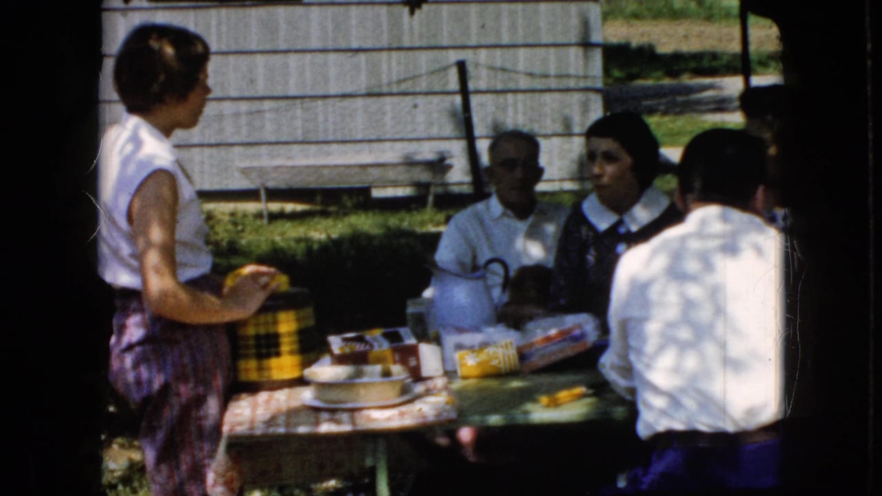 Adults gathered around an outdoor picnic table as a woman stands beside food and a checkered container.