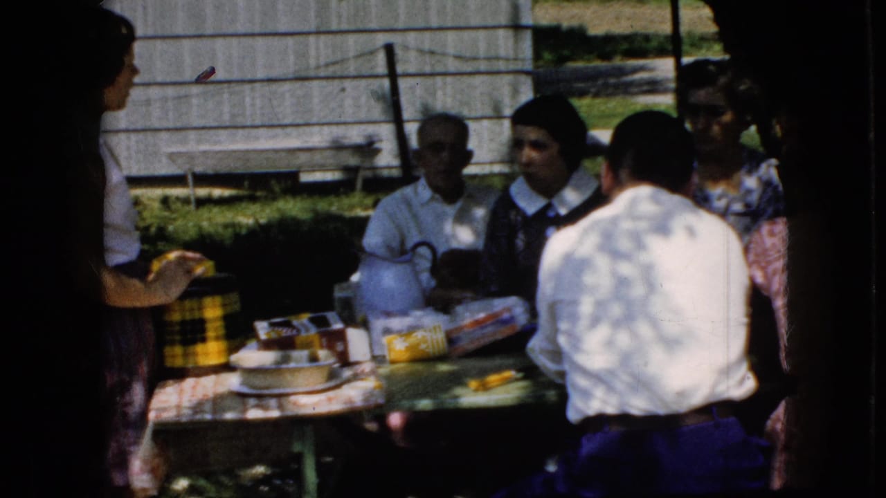 1969 California home-movie frame of people gathered at an outdoor picnic table with food packaging, plates, and a white pitcher or kettle in front of a wooden outbuilding.