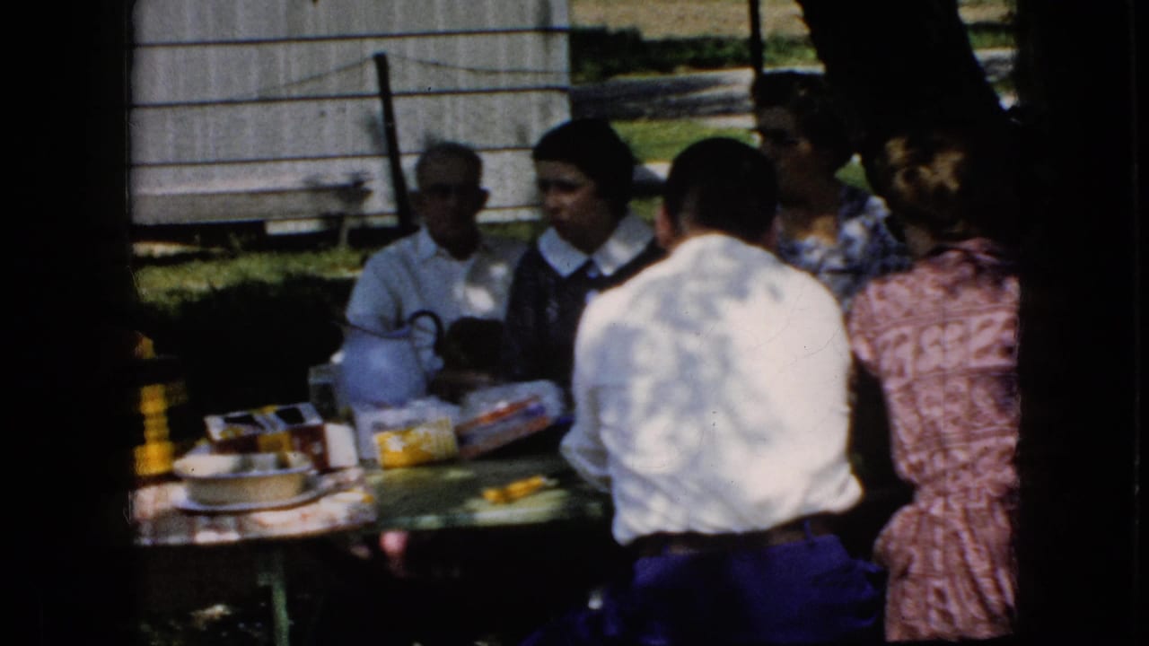1969 California home-movie frame showing adults seated around a picnic table with visible packaged food and a woman in a pink patterned dress near the right edge.