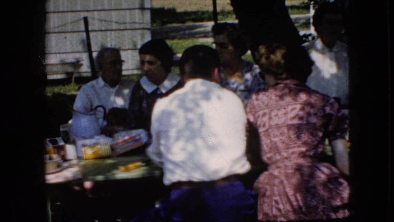 1969 California home-movie frame showing several adults around an outdoor picnic table, with food packages, tableware, and a wooden outbuilding behind them.