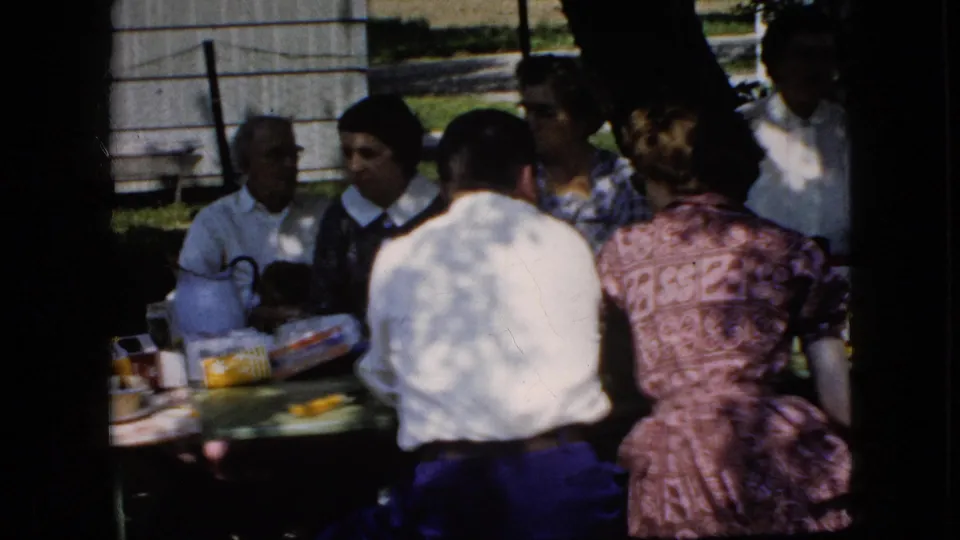 1969 California home-movie frame showing several adults around an outdoor picnic table, with food packages, tableware, and a wooden outbuilding behind them.