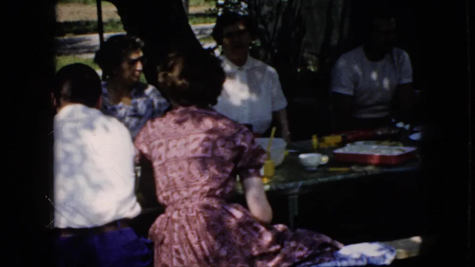 1969 California 8mm frame showing a shaded outdoor meal with adults seated closely around a table set with cups, bowls, trays, and snacks.