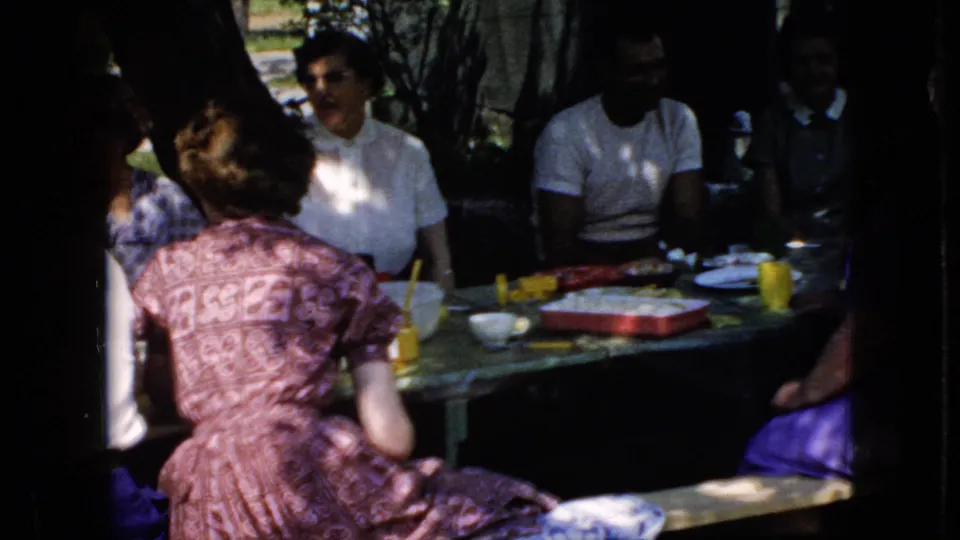 1969 California home-movie frame showing adults at a shaded picnic table, with food trays, yellow cups, and casual conversation visible in the darkened exposure.