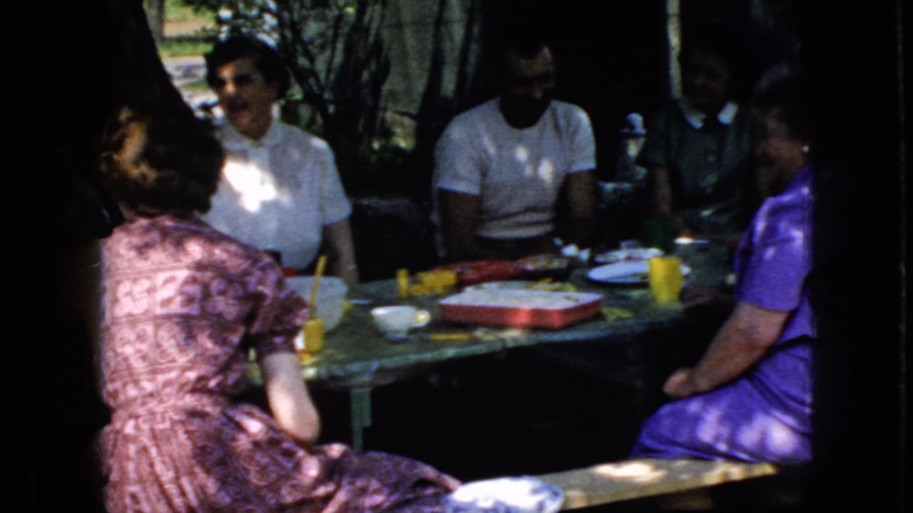 1969 California home-movie frame showing adults gathered around a shaded outdoor table with colorful cups, plates, and containers.