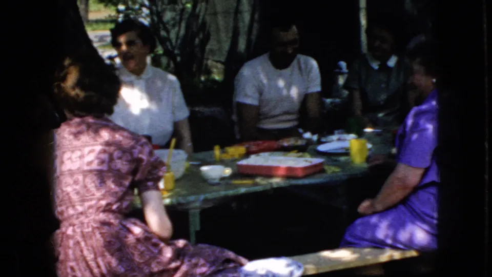 1969 California home-movie frame showing adults gathered around a shaded outdoor table with colorful cups, plates, and containers.