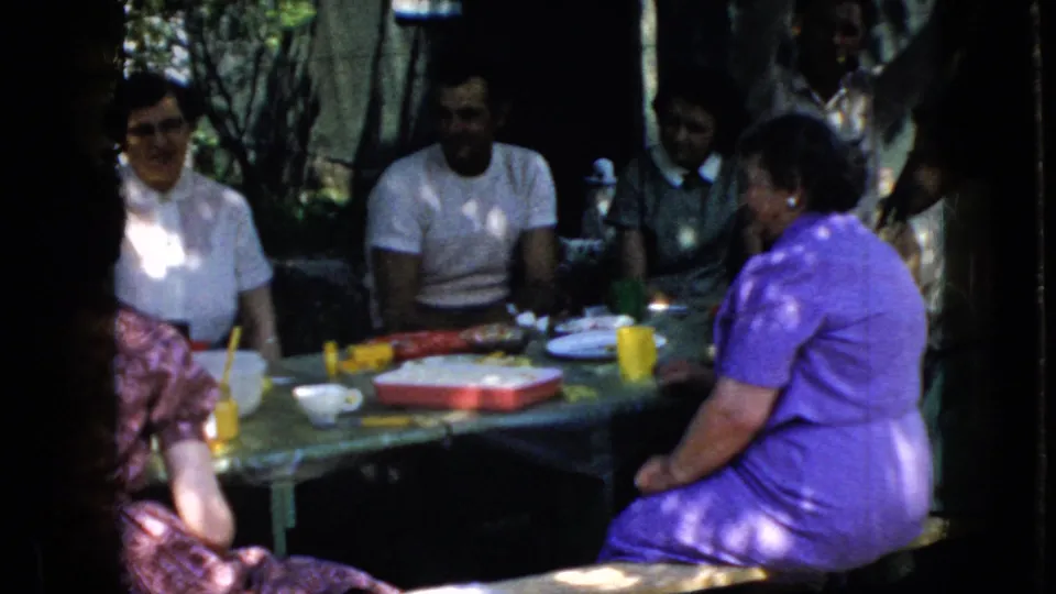 1969 California home-movie frame of adults seated around an outdoor meal table beneath trees, with dappled sunlight across clothing and tableware.