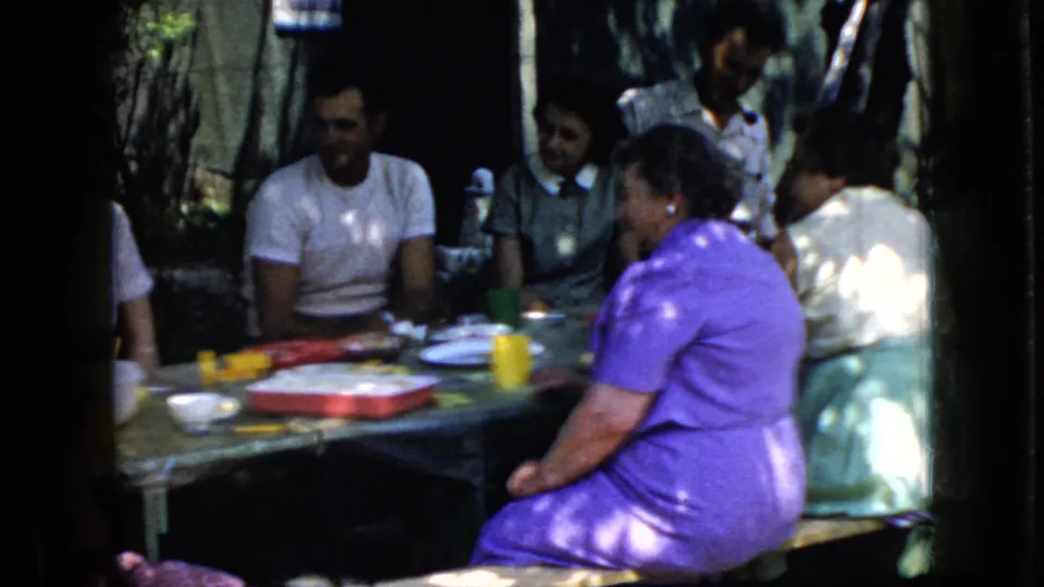 1969 California home-movie frame showing a small group of adults at a shaded picnic table with food containers, cups, and plates visible.