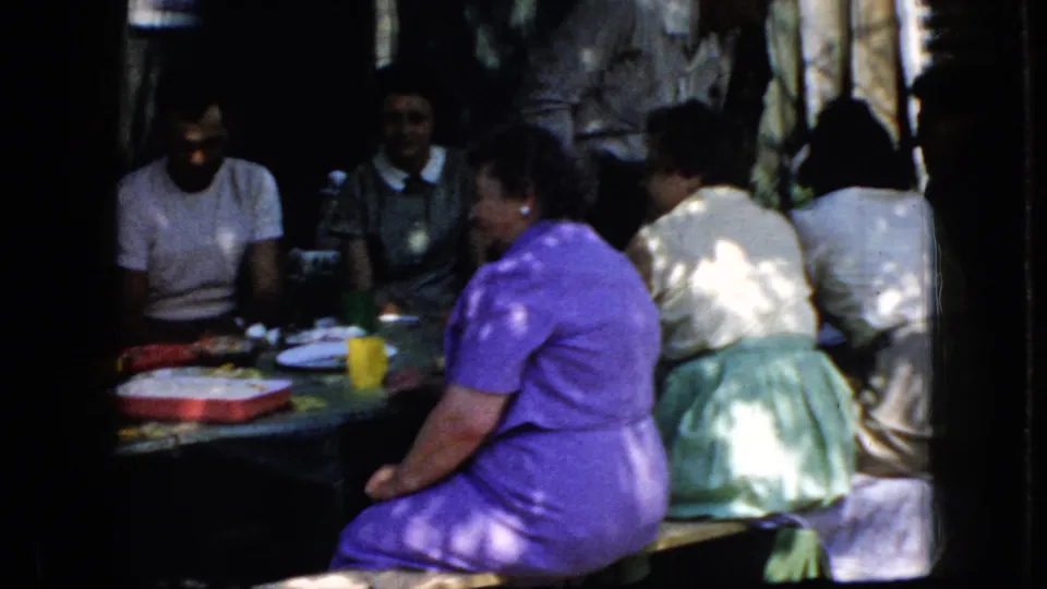 1969 California home-movie frame of adults sitting closely around a wooded picnic table with cups, plates, and containers on the table.