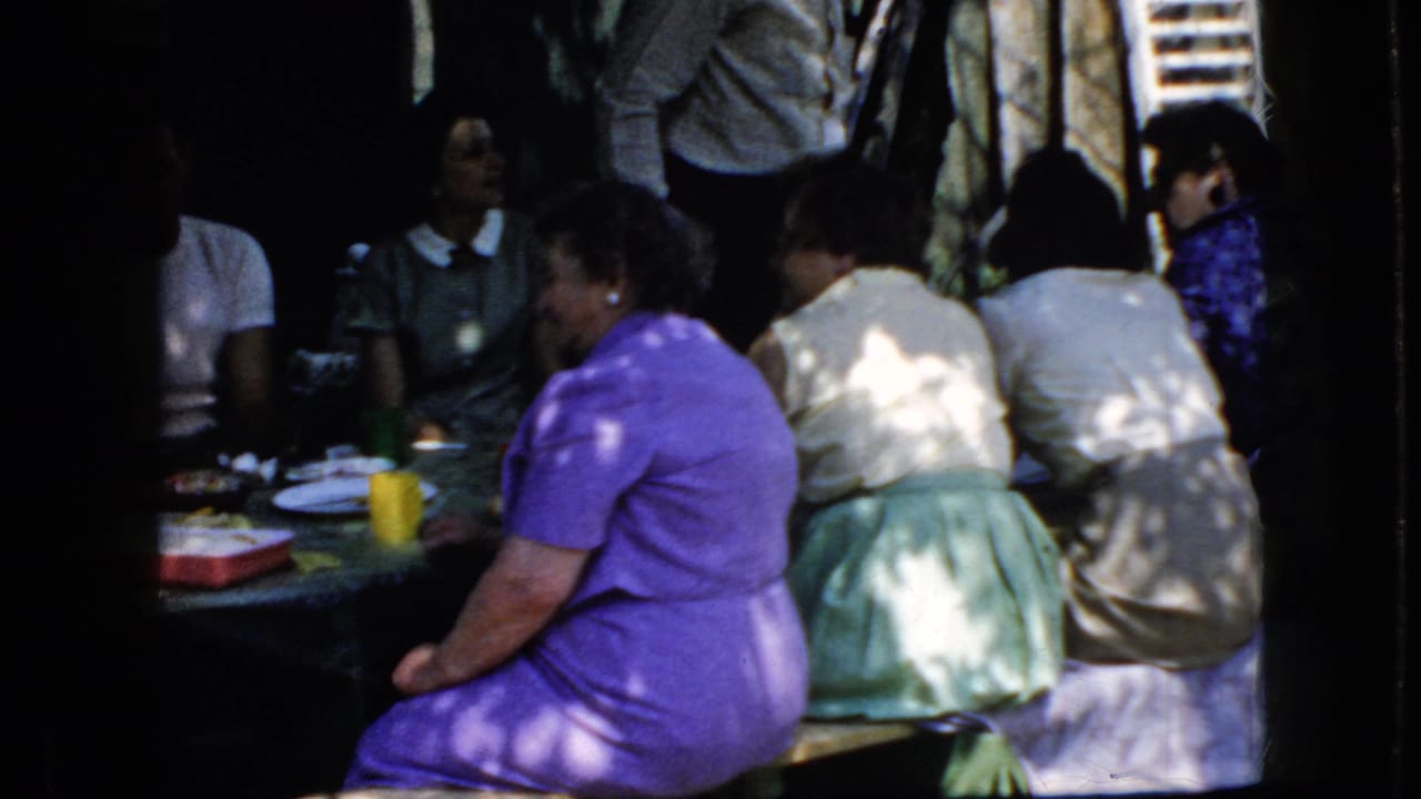 California 1969 home-movie frame showing adult women and other attendees seated at an outdoor picnic table in mottled sunlight.